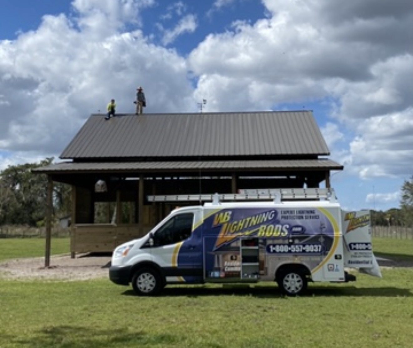 WB Lightning Rods van parked in front of an open pole barn pavilion with two crew members on the metal roof